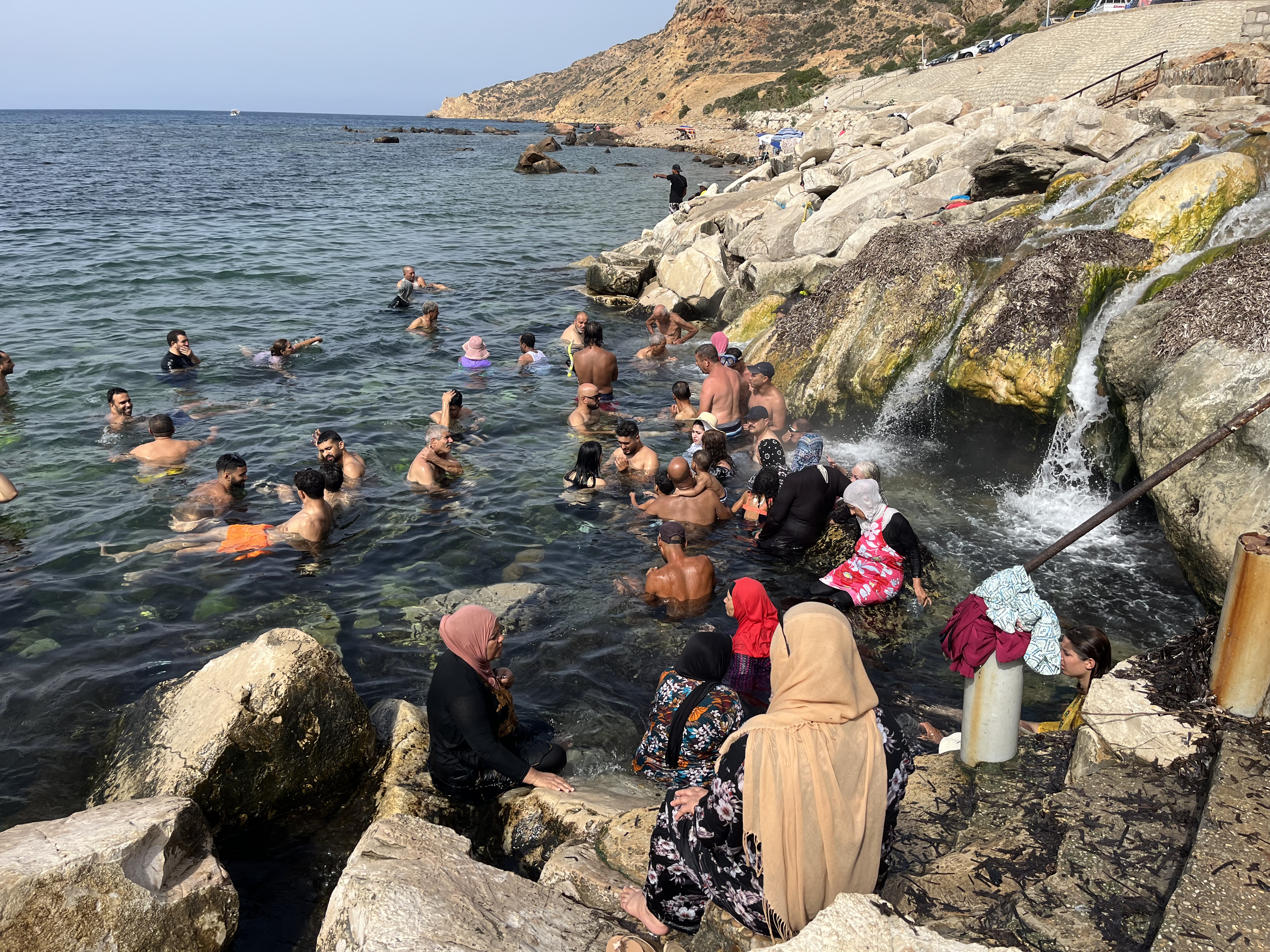 A crowd of people swimming in the sea, next to a waterfall